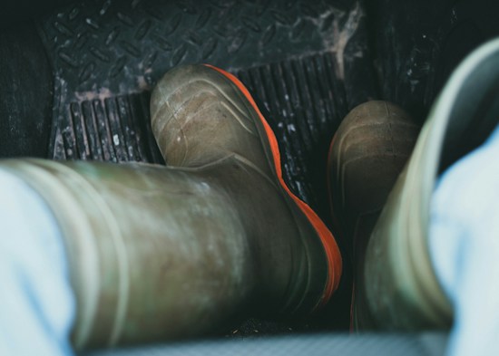 View of a pair of worn work boots with orange soles, placed on the rubber floor mat of a vehicle interior. The floor mat has a rugged texture, and the surrounding floor area seems to be somewhat dirty, indicating use in muddy or rough environments.