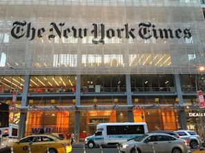 A modern glass building with the New York Times logo prominently displayed across the upper facade. The lower levels of the building show large windows revealing office interiors with bright lighting and visible desks. Several vehicles, including a yellow taxi, a white express service bus, and a dark sedan, are on the street in front of the building.