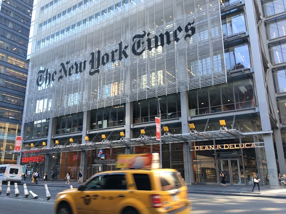 A busy urban street scene in front of a modern glass and steel building with 'The New York Times' signage prominently displayed. Several people are walking on the sidewalk. A yellow taxi is passing by in the foreground and storefronts for 'Dean & DeLuca' and 'Schnippers' are visible.