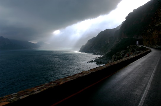 A coastal road runs alongside steep cliffs, with dramatic clouds overhead and sunlight breaking through. The ocean stretches towards the horizon, with waves crashing against the rocks.