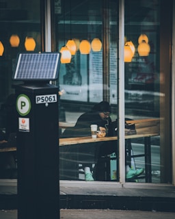 A person sitting inside a cafe, visible through the large glass windows, appears to be working or studying on a laptop. The person is wearing a dark coat and bright turquoise sneakers. Several warm, yellow pendant lights hang from the ceiling, creating a cozy atmosphere. Outside the cafe, there is a parking meter in the foreground.