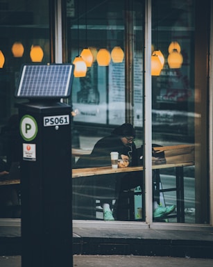 A person sitting inside a cafe, visible through the large glass windows, appears to be working or studying on a laptop. The person is wearing a dark coat and bright turquoise sneakers. Several warm, yellow pendant lights hang from the ceiling, creating a cozy atmosphere. Outside the cafe, there is a parking meter in the foreground.