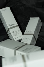 Several white boxes with a minimalist design are scattered on a dark surface. The boxes are branded with 'The Ordinary' and contain skincare products such as Caffeine Solution 5% + EGCG and 100% Organic Cold-Pressed Moroccan Argan Oil. The boxes contain details about each product, including usage instructions and quantities.