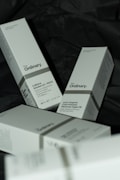 Several white boxes with a minimalist design are scattered on a dark surface. The boxes are branded with 'The Ordinary' and contain skincare products such as Caffeine Solution 5% + EGCG and 100% Organic Cold-Pressed Moroccan Argan Oil. The boxes contain details about each product, including usage instructions and quantities.
