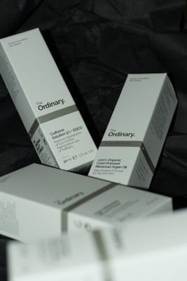 Several white boxes with a minimalist design are scattered on a dark surface. The boxes are branded with 'The Ordinary' and contain skincare products such as Caffeine Solution 5% + EGCG and 100% Organic Cold-Pressed Moroccan Argan Oil. The boxes contain details about each product, including usage instructions and quantities.