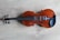A violin is laid flat on a light wooden surface. The violin has a rich, brown varnish that highlights its natural wood grain, complemented by black tuning pegs and fingerboard. The subtle textures and patterns of the wooden background enhance the elegant simplicity of the instrument.