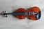 A violin is laid flat on a light wooden surface. The violin has a rich, brown varnish that highlights its natural wood grain, complemented by black tuning pegs and fingerboard. The subtle textures and patterns of the wooden background enhance the elegant simplicity of the instrument.
