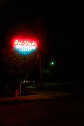 A neon sign glows brightly in the dark, featuring the words 'Bel-Vista Healthcare Center 5001'. The sign is illuminated with red and blue light, casting its glow on the surrounding area. In the background, a tree and a streetlight are visible, creating a moody atmosphere.
