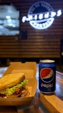 A juicy cheeseburger with lettuce is placed inside a takeout box next to a can of Pepsi on a wooden table. The background features a blurry sign with white neon lights.