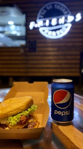 A juicy cheeseburger with lettuce is placed inside a takeout box next to a can of Pepsi on a wooden table. The background features a blurry sign with white neon lights.