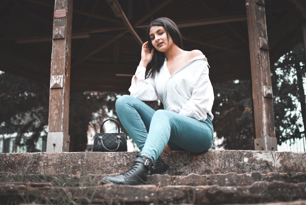 woman in white long sleeve shirt and blue denim jeans sitting on brown concrete stairs