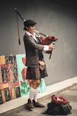 A person wearing traditional Scottish attire, including a kilt and hat, is playing bagpipes. They stand on a sidewalk next to a wall displaying several colorful paintings with floral and abstract designs. A backpack filled with items, possibly for collecting tips, rests on the ground nearby.