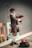 A person wearing traditional Scottish attire, including a kilt and hat, is playing bagpipes. They stand on a sidewalk next to a wall displaying several colorful paintings with floral and abstract designs. A backpack filled with items, possibly for collecting tips, rests on the ground nearby.