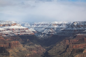 A vast expanse of the Grand Canyon, partially covered with a light dusting of snow. The rock formations reveal layers of red and brown hues, creating a striking contrast with the white snow. The landscape showcases deep valleys and rugged cliffs with a cloudy sky above.