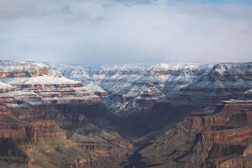 A vast expanse of the Grand Canyon, partially covered with a light dusting of snow. The rock formations reveal layers of red and brown hues, creating a striking contrast with the white snow. The landscape showcases deep valleys and rugged cliffs with a cloudy sky above.