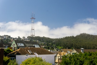 A residential area with houses in the foreground and a large hill covered in trees in the background. A tall communication tower stands prominently on the hill, under a partly cloudy sky.