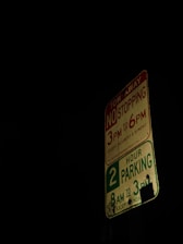 A parking sign is illuminated against a dark background. It displays restrictions including no stopping from 3 PM to 6 PM, and two-hour parking from 8 AM to 3 PM, with certain exceptions.