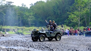 A group of people is enjoying an outdoor adventure in jeeps on a rocky terrain surrounded by lush green forest. One jeep is in the foreground with passengers expressing excitement and a driver focusing on the path ahead. In the background, more people and vehicles are visible, indicating a larger group or event.