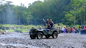 A group of people is enjoying an outdoor adventure in jeeps on a rocky terrain surrounded by lush green forest. One jeep is in the foreground with passengers expressing excitement and a driver focusing on the path ahead. In the background, more people and vehicles are visible, indicating a larger group or event.