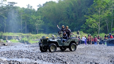 A group of people is enjoying an outdoor adventure in jeeps on a rocky terrain surrounded by lush green forest. One jeep is in the foreground with passengers expressing excitement and a driver focusing on the path ahead. In the background, more people and vehicles are visible, indicating a larger group or event.