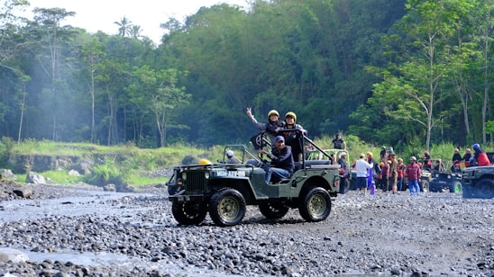 A group of people is enjoying an outdoor adventure in jeeps on a rocky terrain surrounded by lush green forest. One jeep is in the foreground with passengers expressing excitement and a driver focusing on the path ahead. In the background, more people and vehicles are visible, indicating a larger group or event.
