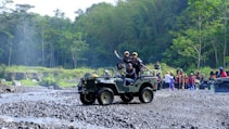 A group of people is enjoying an outdoor adventure in jeeps on a rocky terrain surrounded by lush green forest. One jeep is in the foreground with passengers expressing excitement and a driver focusing on the path ahead. In the background, more people and vehicles are visible, indicating a larger group or event.