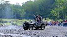 A group of people is enjoying an outdoor adventure in jeeps on a rocky terrain surrounded by lush green forest. One jeep is in the foreground with passengers expressing excitement and a driver focusing on the path ahead. In the background, more people and vehicles are visible, indicating a larger group or event.
