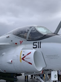 A military jet aircraft is parked, with the cockpit canopy closed. The fuselage features prominent markings and warnings such as 'JET INTAKE' and 'DANGER.' The jet is painted in a light color, likely gray or white, and the number '511' is visible near the cockpit. The background shows a cloudy sky.