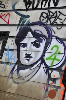 A mural or graffiti on a wall featuring a stylized black and white portrait of a person, surrounded by colorful abstract shapes and letters. The image includes bold outlines and a mix of spray-painted text and symbols.