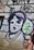 A mural or graffiti on a wall featuring a stylized black and white portrait of a person, surrounded by colorful abstract shapes and letters. The image includes bold outlines and a mix of spray-painted text and symbols.