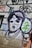 A mural or graffiti on a wall featuring a stylized black and white portrait of a person, surrounded by colorful abstract shapes and letters. The image includes bold outlines and a mix of spray-painted text and symbols.