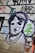 A mural or graffiti on a wall featuring a stylized black and white portrait of a person, surrounded by colorful abstract shapes and letters. The image includes bold outlines and a mix of spray-painted text and symbols.