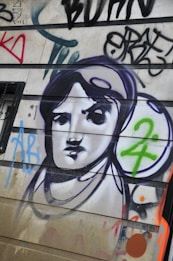 A mural or graffiti on a wall featuring a stylized black and white portrait of a person, surrounded by colorful abstract shapes and letters. The image includes bold outlines and a mix of spray-painted text and symbols.