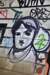 A mural or graffiti on a wall featuring a stylized black and white portrait of a person, surrounded by colorful abstract shapes and letters. The image includes bold outlines and a mix of spray-painted text and symbols.