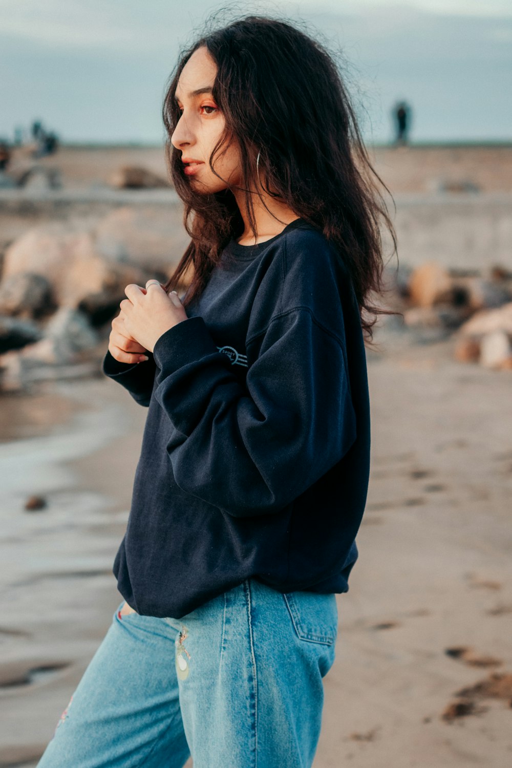 woman in black jacket and blue denim jeans standing on beach during daytime