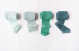Four neatly rolled strips of fabric in different shades of green are placed side by side on a white surface. The fabric strips have frayed edges, giving them a textured, soft appearance. They gradually transition from a light mint green on the left to a deeper teal on the right.