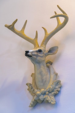 A wall-mounted sculpture of a deer's head with large, intricate antlers, featuring realistic detailing. The sculpture is mounted on an ornamental base that includes decorative floral elements.