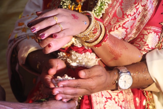 Intricate henna designs decorate a woman's hands, adorned with gold rings and bangles. The vibrant red and gold sari adds to the festive atmosphere, with flowers and confetti sprinkled around the wrist. A watch is visible on an accompanying hand, suggesting a cultural or celebratory event.