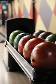 A row of bowling balls of various colors are neatly lined up on a ball rack. The background features a colorful geometric wall design, adding to the vibrant atmosphere typical of a bowling alley.