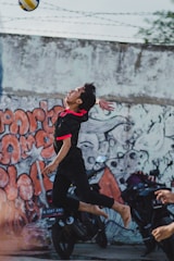 A person is leaping barefoot to hit a volleyball in an urban setting. The backdrop features a colorful graffiti-covered wall. Motorcycles are parked along the wall, adding to the urban vibe. The player wears a black sports outfit with bright red accents.