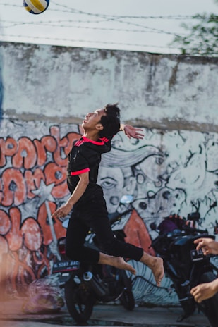 A person is leaping barefoot to hit a volleyball in an urban setting. The backdrop features a colorful graffiti-covered wall. Motorcycles are parked along the wall, adding to the urban vibe. The player wears a black sports outfit with bright red accents.