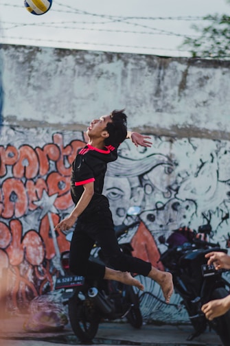 A person is leaping barefoot to hit a volleyball in an urban setting. The backdrop features a colorful graffiti-covered wall. Motorcycles are parked along the wall, adding to the urban vibe. The player wears a black sports outfit with bright red accents.