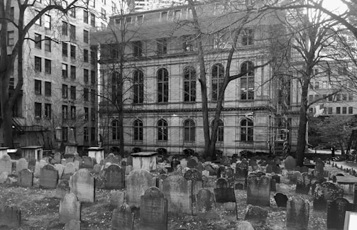 A historic cemetery is surrounded by old stone buildings, with numerous weathered tombstones scattered across the grassy area. Bare trees stand amid the headstones, their branches reaching upwards. The architecture of the surrounding buildings features classic, arched windows and intricate details. Scaffolding can be seen on one of the buildings, indicating ongoing maintenance or restoration work.