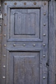 A weathered wooden door featuring a geometric pattern with two large rectangular panels and a smaller central square panel. The surface is adorned with metallic round studs, and the wood exhibits signs of aging with a dark, muted finish. There are decorative vertical grooves on the right side frame.