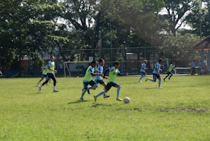 A group of young athletes playing soccer on a grassy field, surrounded by trees and a fence. Players are wearing matching blue uniforms, and a few are in yellow pinnies. The scene is set under a clear sky with ample sunlight.