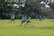 A group of young athletes playing soccer on a grassy field, surrounded by trees and a fence. Players are wearing matching blue uniforms, and a few are in yellow pinnies. The scene is set under a clear sky with ample sunlight.