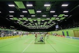 A sports hall with bright green flooring and ceiling panels. There is a game in progress with players moving quickly, suggesting a fast-paced sport like floorball or indoor hockey. Advertising banners line the walls, and a scoreboard is visible in the background.