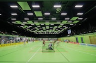 A sports hall with bright green flooring and ceiling panels. There is a game in progress with players moving quickly, suggesting a fast-paced sport like floorball or indoor hockey. Advertising banners line the walls, and a scoreboard is visible in the background.