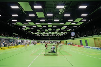A sports hall with bright green flooring and ceiling panels. There is a game in progress with players moving quickly, suggesting a fast-paced sport like floorball or indoor hockey. Advertising banners line the walls, and a scoreboard is visible in the background.