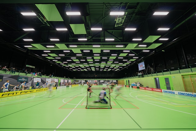 A sports hall with bright green flooring and ceiling panels. There is a game in progress with players moving quickly, suggesting a fast-paced sport like floorball or indoor hockey. Advertising banners line the walls, and a scoreboard is visible in the background.